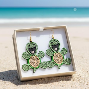 Puede incluir: Un par de pendientes de tortuga verdes con ganchos dorados, presentados en una caja blanca. Los pendientes presentan un diseño de tortuga sonriente con ojos negros y un caparazón marrón claro. El fondo muestra una playa de arena y el océano.