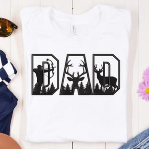 May include: White t-shirt with the word "DAD" in a black, outdoor-themed design. The letter "D" features an archer, the "A" has a deer head, and the final "D" shows a deer silhouette. The design includes trees.