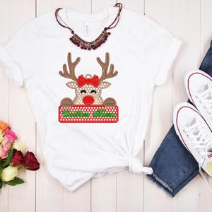 Op de afbeelding: Wit T-shirt met een rendierontwerp, een rode strik en de tekst "Custom Name" op een rood en groen bord. De afbeelding bevat een blauwe jeans, witte sneakers, een ketting en bloemen. Het T-shirt is geknoopt in de taille.