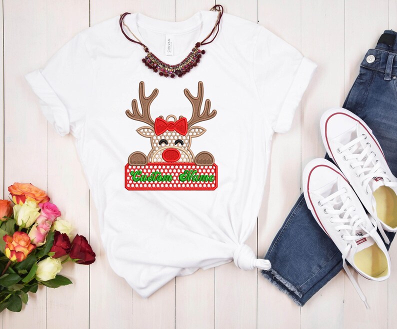Op de afbeelding: Wit T-shirt met een rendierontwerp, een rode strik en de tekst "Custom Name" op een rood en groen bord. De afbeelding bevat een blauwe jeans, witte sneakers, een ketting en bloemen. Het T-shirt is geknoopt in de taille.