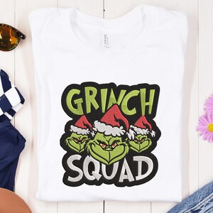 May include: White t-shirt with a Grinch Squad graphic. The graphic features the words "GRINCH SQUAD" in green and white, with three Grinch faces wearing Santa hats. The shirt is folded on a white wooden surface.