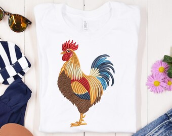 May include: White t-shirt featuring a detailed embroidered rooster design. The rooster has a red comb, yellow beak, and a colorful plumage of red, yellow, blue, and brown. The shirt is a casual wear item.