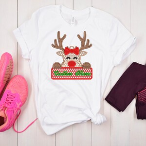 Op de afbeelding: Wit T-shirt met een rendierontwerp. Het rendier heeft bruine geweien, een rode strik en een rood en groen bord met de tekst "Custom Name". Het rendier heeft een stippenpatroon. Roze sneakers en bordeauxrode leggings zijn ook zichtbaar.