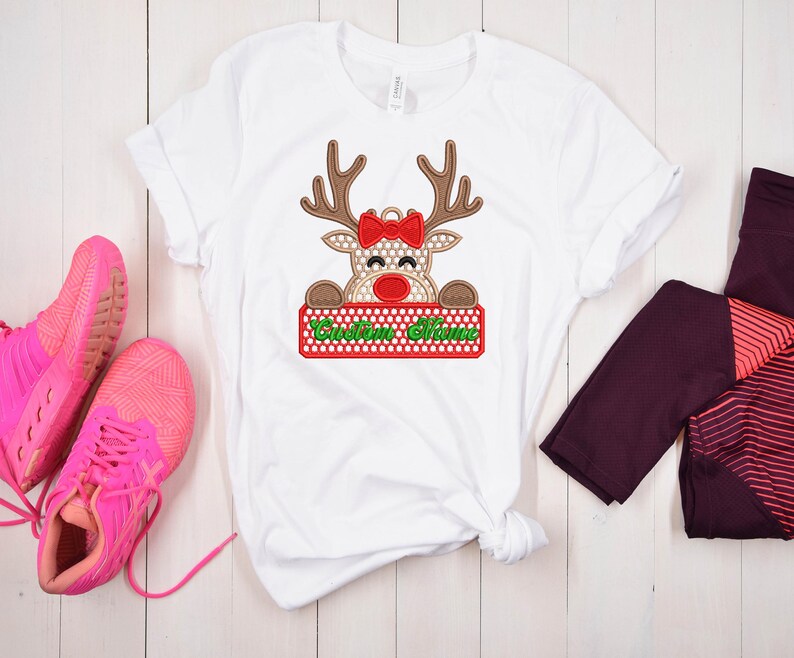 Op de afbeelding: Wit T-shirt met een rendierontwerp. Het rendier heeft bruine geweien, een rode strik en een rood en groen bord met de tekst "Custom Name". Het rendier heeft een stippenpatroon. Roze sneakers en bordeauxrode leggings zijn ook zichtbaar.