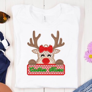 Op de afbeelding: Wit T-shirt met een rendierontwerp. Het rendier heeft bruine geweien, een rode strik en een rode neus. Het houdt een rood bord met de tekst "Custom Name" in groen. Een feestelijk kerstontwerp.