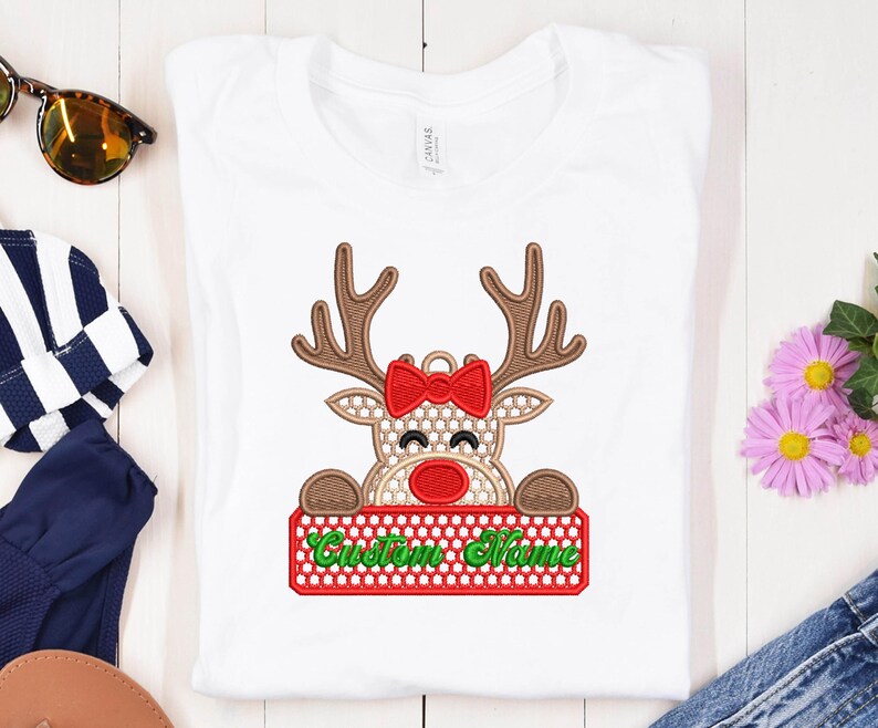 Op de afbeelding: Wit T-shirt met een rendierontwerp. Het rendier heeft bruine geweien, een rode strik en een rode neus. Het houdt een rood bord met de tekst "Custom Name" in groen. Een feestelijk kerstontwerp.