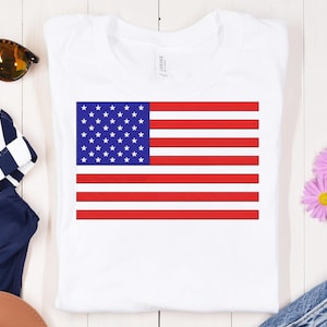 May include: White t-shirt with an embroidered American flag design. The flag shows red and white stripes and a blue square with white stars. The t-shirt is a classic crew neck style.