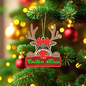 Op de afbeelding: Geborduurde kerstversiering met een rendierontwerp, een rode strik en een rood bord met de tekst "Custom Name" in het groen. De versiering hangt aan een kerstboom met rode ballen en gele lampjes.