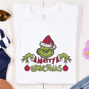 Può includere: T-shirt bianca con un personaggio verde del Grinch con un cappello da Babbo Natale. Il Grinch tiene due ornamenti rossi, con le parole "Merry Grinchmas" in rosso e verde.