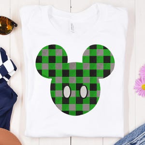 Könnte beinhalten: Weißes T-Shirt mit einem grün, schwarz und grau karierten Mickey Mouse-Design. Das Design zeigt die ikonischen Ohren und das Gesicht der Maus. Das T-Shirt ist gefaltet und auf einer weißen Holzoberfläche ausgestellt.