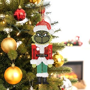 May include: A Christmas tree ornament featuring a green character with a Santa hat and red coat. The ornament has a black belt, green pants, and black boots. It is hanging on a decorated Christmas tree with red and gold ornaments.