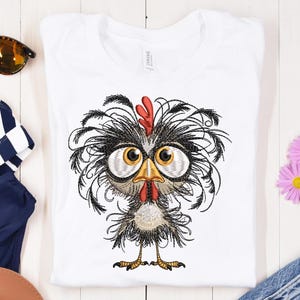 May include: White t-shirt featuring an embroidered cartoon chicken with large eyes, a red comb, and a yellow beak. The chicken has black and gray feathers. The shirt is made of a soft material.