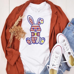 May include: A white t-shirt with a cartoon bunny design in red, white, and blue stripes, with star accents and a yellow bow tie. The bunny is sitting. The shirt is styled with a rust-coloured blazer, blue jeans, and white trainers.