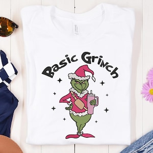 May include: White t-shirt with a cartoon Grinch character wearing a Santa hat and pink outfit, holding a pink drink. The text "Basic Grinch" is above the character in black. Four black stars surround the Grinch.