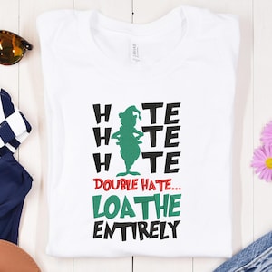 May include: A white t-shirt with a green cartoon character and the text "HATE HATE HATE DOUBLE HATE... LOATHE ENTIRELY" in black and green. The shirt is folded and displayed on a light-coloured wooden surface.