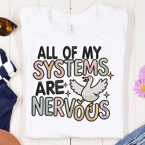 Peut inclure: T-shirt blanc avec l'inscription "ALL OF MY SYSTEMS ARE NERVOUS" dans une police de caractères colorée et ludique. Un canard de dessin animé aux ailes déployées est présenté en dessous du texte, avec des étoiles. Le t-shirt est présenté sur une surface en bois clair.