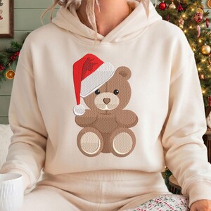 May include: A cream-coloured hooded sweatshirt featuring an embroidered design of a brown teddy bear wearing a red and white Santa hat. The bear has a light brown muzzle and circular light brown patches on its feet, centred on the chest.