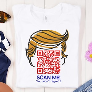 May include: White t-shirt with a cartoon illustration of a hairstyle and a red QR code. The text "SCAN ME! You won't regret it." is printed below the QR code in blue.