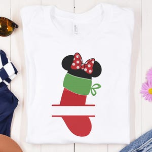 May include: White t-shirt featuring a red and green stocking design with a Minnie Mouse bow and ears. The design includes a blank space for personalization. Sunglasses and other accessories are visible in the background.