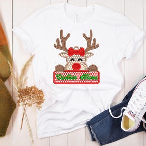 Op de afbeelding: Wit T-shirt met een rendierontwerp met een rode strik, rode neus en bruin gewei. Het rendier houdt een bord vast met "Custom Name" in het groen. Het shirt wordt gecombineerd met een blauwe jeans en witte sneakers.