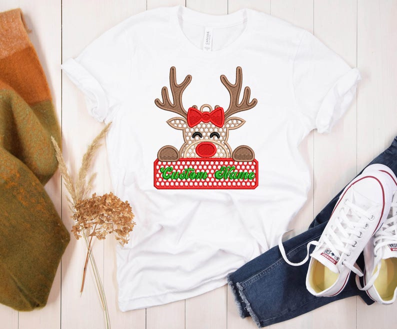 Op de afbeelding: Wit T-shirt met een rendierontwerp met een rode strik, rode neus en bruin gewei. Het rendier houdt een bord vast met "Custom Name" in het groen. Het shirt wordt gecombineerd met een blauwe jeans en witte sneakers.