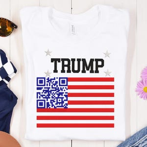 Peut inclure: T-shirt blanc avec un motif de drapeau américain rouge, blanc et bleu. Le drapeau comporte un code QR dans le coin supérieur gauche et le mot "TRUMP" en lettres noires au-dessus du drapeau.