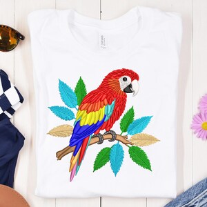 Puede incluir: Camiseta blanca con un diseño bordado de loro colorido. El loro tiene plumas rojas, amarillas, azules y verdes, posado en una rama con hojas azules, verdes y doradas. Un diseño vibrante y elegante.