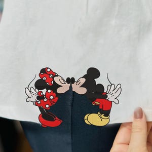 May include: White t-shirt with a red and black cartoon image of Mickey Mouse and Minnie Mouse kissing.