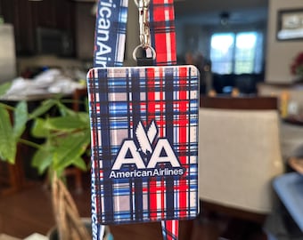 May include: A rectangular ID badge holder with a red, white, and blue plaid pattern featuring the American Airlines logo and text. It is attached to a matching fabric lanyard with a metal swivel hook and a black plastic safety breakaway clasp.