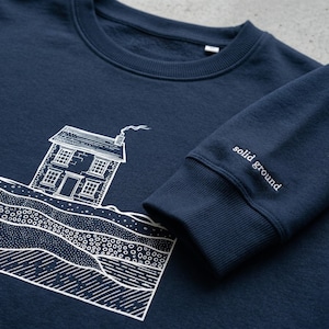 Könnte beinhalten: Marineblaues Sweatshirt mit weißer Illustration eines Hauses und geologischer Schichten. Am Ärmelbund befindet sich der gestickte Schriftzug "solid ground". Das Sweatshirt ist aus weichem Material.