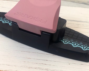 May include: A Fiskars paper punch in a combination of black and pink. The punch has a pink handle with the brand name Fiskars embossed on it. The base is black with a decorative teal design along the edge. The punch is designed for crafting.