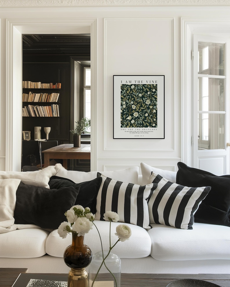 May include: A living room scene featuring a white sofa adorned with black and white striped and solid black pillows. A framed art print on the wall reads "I AM THE VINE" above a floral pattern. A vase of white flowers sits on the coffee table.