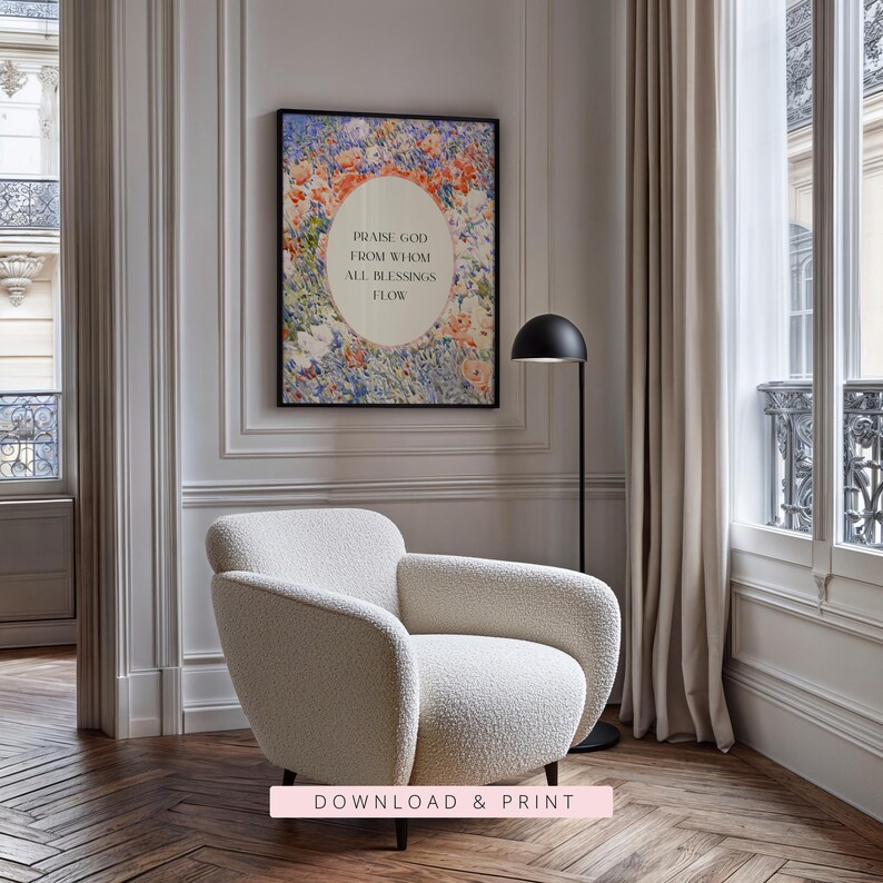 May include: A cream-colored textured armchair sits in front of a wall-mounted framed print featuring a floral border and the text "Praise God from whom all blessings flow" centered within an oval frame, accompanied by a black floor lamp.