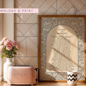 K&ouml;nnte beinhalten: Ein gerahmter Druck mit dem Text der Litanei der Demut, zentriert in einem beigen Bogenfeld, umgeben von einem blau-wei&szlig;en Blumenmuster. Der Druck befindet sich in einem Holzrahmen neben einem rosa Toaster und einer wei&szlig;en Tasse mit Zickzackmuster.