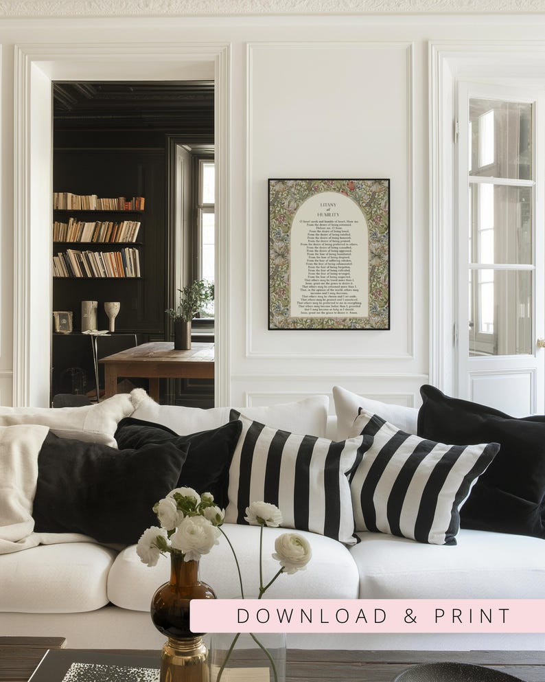 May include: A framed wall art print featuring the text Litany of Humility surrounded by a colourful floral and botanical border. The print is displayed on a white wall above a white sofa with black and white striped and solid black throw pillows.