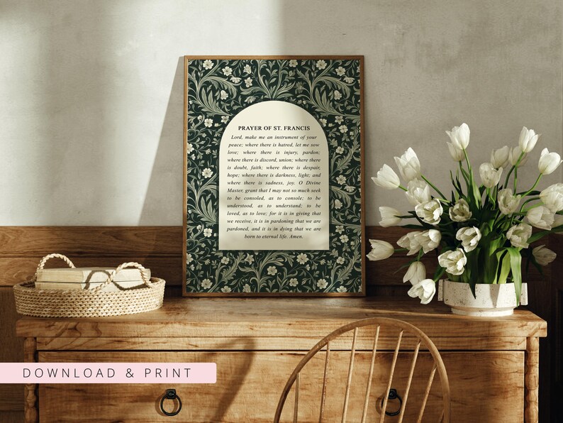 May include: A framed print of the Prayer of St. Francis, featuring a dark green floral pattern border. The text is centered on a cream-colored background. A basket and a vase of white tulips sit on a wooden dresser.