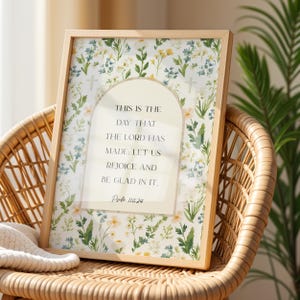 May include: A framed print with a light wood frame. The print features a floral pattern with white, yellow, and green flowers. The text reads: "THIS IS THE DAY THAT THE LORD HAS MADE: LET US REJOICE AND BE GLAD IN IT." Psalm 118:24.