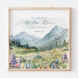 May include: A square framed wall art print featuring a watercolor landscape of mountains, pine trees, and a wildflower meadow. The text reads: HE IS NOT HERE: He has Risen JUST AS HE SAID. Matthew 28:6. The colors include blue, green, and yellow.