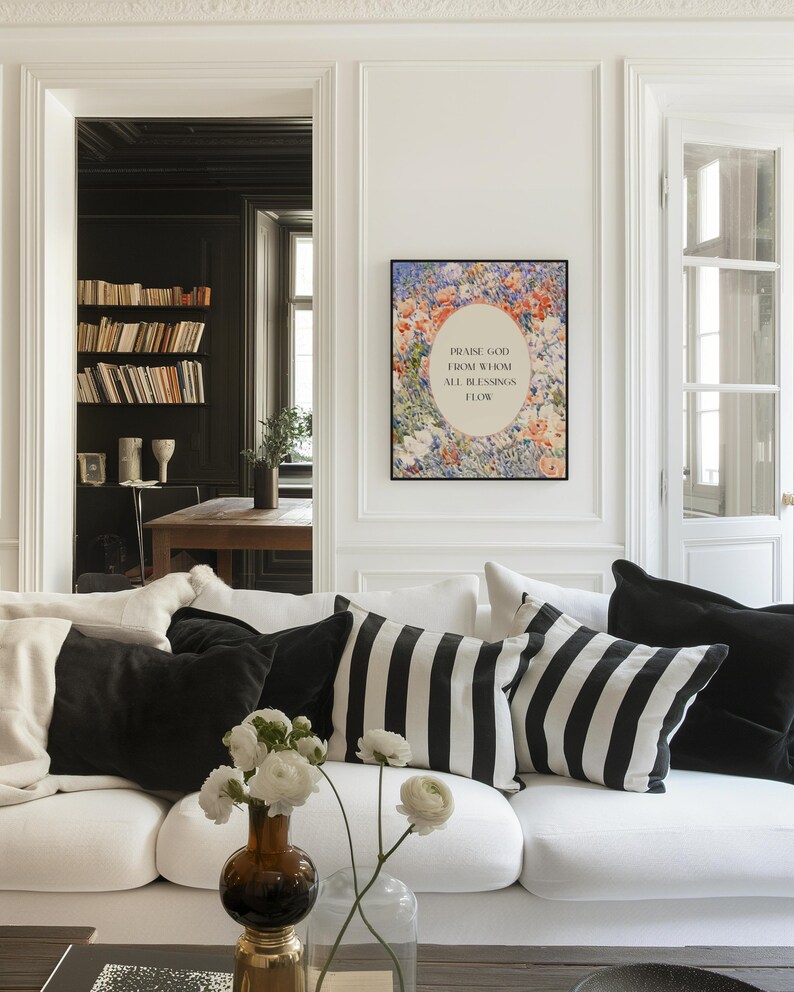 May include: A rectangular wall art print in a black frame featuring a floral border surrounding an oval center with the text "PRAISE GOD FROM WHOM ALL BLESSINGS FLOW" in black lettering, displayed on a white wall above a white sofa with pillows.