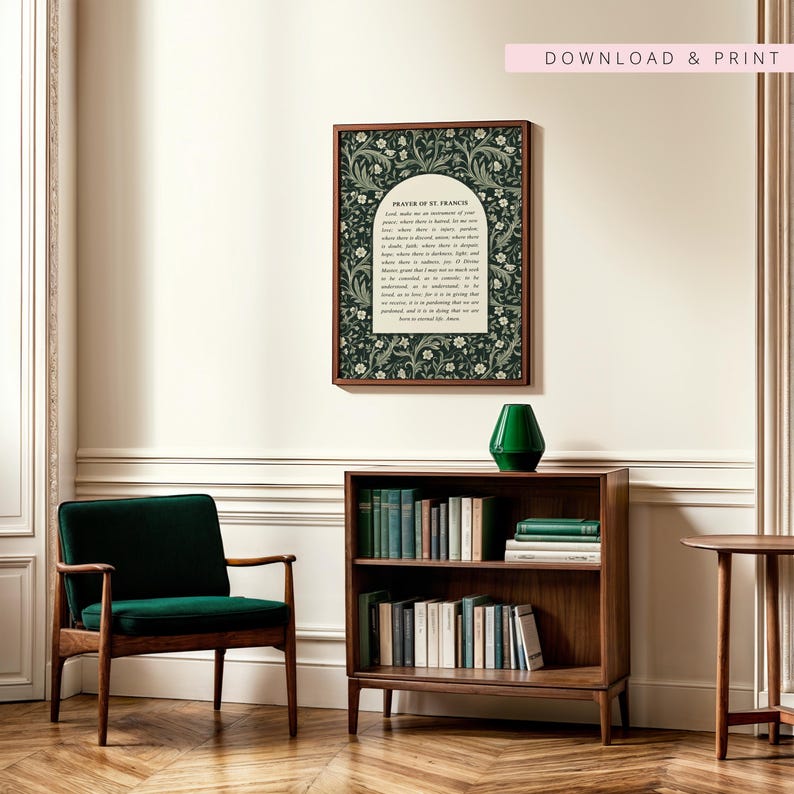 May include: A framed print featuring the "Prayer of St. Francis" with a dark green floral design. The print hangs above a wooden bookshelf filled with books and a green vase. A green armchair and a small side table are also visible.