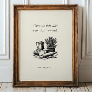 以下が含まれることがあります： Give us this day our daily breadおよびMatthew 6:11というテキストが記された額装済みのウォールアート。中央のイラストは、パン、ピッチャー、麦の入ったバスケット、麺棒を描いた白黒の線画です。