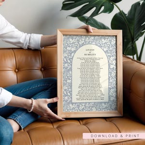 K&ouml;nnte beinhalten: Ein gerahmter Druck mit dem Text der Litanei der Demut, zentriert in einem cremefarbenen Bogenfeld, umgeben von einem hellblauen und wei&szlig;en Blumenmuster. Das Kunstwerk befindet sich in einem hellen Holzrahmen mit einem schlichten Rand.