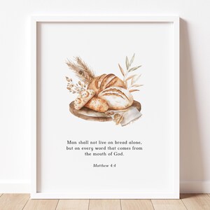 Op de afbeelding: Waterverfprint met verschillende soorten brood, waaronder een brood en broodjes, gerangschikt op een houten plank met tarwe- en bladaccenten. De print bevat de tekst "Man shall not live on bread alone, but on every word that comes from the mouth of God. Matthew 4:4."