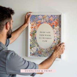 May include: A rectangular white frame containing a print with a colorful floral background of red, blue, and white flowers. In the center, an oval shape features the black text: PRAISE GOD FROM WHOM ALL BLESSINGS FLOW. The print is displayed on a wall.