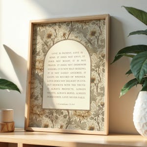 May include: A framed print with the text "Love is Patient" from 1 Corinthians 13:4-8. The print has a light wood frame and a floral patterned background in beige and grey. The text is in a cream-coloured arch.