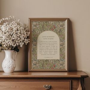 May include: A framed print with the text "LOVE IS PATIENT LOVE IS KIND" in a gold frame. The print has a floral design in green, red, and cream colours. A white vase with white flowers is on the left.