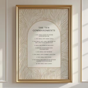 Op de afbeelding: Een ingelijste print met "De Tien Geboden" in een decoratieve boog. De tekst is zwart op een crèmekleurige achtergrond met een beige, wervelend patroon. De goudkleurige lijst voegt elegantie toe aan het religieuze kunstwerk.