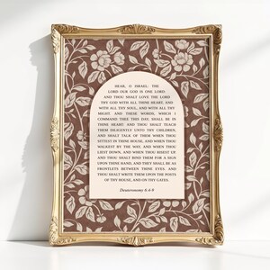 Op de afbeelding: Een goudkleurige, sierlijke lijst met een print in een bruin en crèmekleurig bloemenpatroon. In het midden bevindt zich een crèmekleurig boogpaneel met de zwarte tekst van Deuteronomium 6:4-9, beginnend met Hear, O Israel: The Lord our God is one Lord.