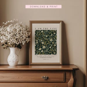 May include: A framed art print with the text "I AM THE VINE" and "YOU ARE THE BRANCHES" over a dark green floral pattern. The print is in a gold frame and sits on a wooden dresser. A white vase with white flowers is to the left.