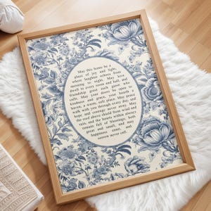 Op de afbeelding: Een rechthoekig ingelijst wandkunstwerk met een crèmekleurige achtergrond en een blauw bloemenpatroon. In het midden bevat een ovaal de tekst: May this home be a place of joy and light, where laughter echoes from morning to night.